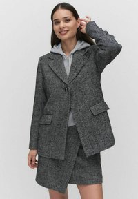 Blazer med houndstooth-mönster i svart och vitt, med en enkel knappstängning, två framfickor och en avslappnad passform över en grå hoodie.