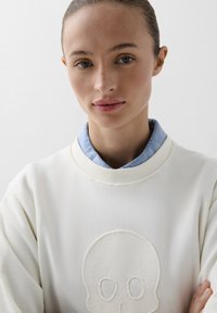 Weißer Sweatshirt mit einem strukturierten Schädel-Logo auf der Vorderseite. Getragen über einem hellblauen Jeanshemd mit Kragen. Glatter Stoff, entspannter Schnitt.