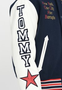 Chaqueta varsity de lana azul marino con mangas de cuero blanco. Presenta "TOMMY" en letras en negrita, una estrella roja bordada y puños acanalados a rayas.