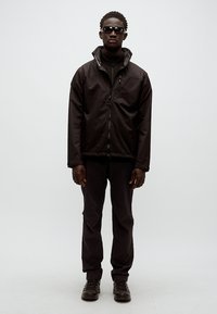 Chaqueta impermeable negra con cuello alto y cierre de cremallera, combinada con pantalones negros a juego y zapatillas oscuras. Modelo con gafas de sol.