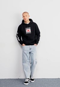 Sudadera negra con un diseño de logo en el frente, jeans baggy de color azul claro y zapatillas negras con acentos blancos. De pie contra una pared lisa.