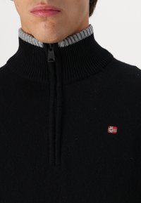 Czarny sweter z dzianiny z wysokim kołnierzem, szarym ściągaczem i centralnym suwakiem. Posiada małe logo norweskiej flagi w kolorach czerwonym i białym.