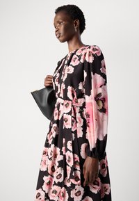 Vestido floral en negro con grandes flores rosas, mangas largas con pliegues y una cintura ajustada. La modelo sostiene un pequeño bolso negro.