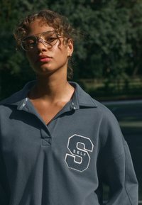 Polo gris con cuello, presenta un "S" bordado texturizado y "SELF". Tejido ligero con un ajuste relajado.
