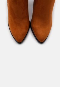 Stivaletti in suede marrone con punta affilata e tacco a stiletto alto, caratterizzati da una texture liscia e un design elegante. Le suole nere contrastano con la parte superiore.