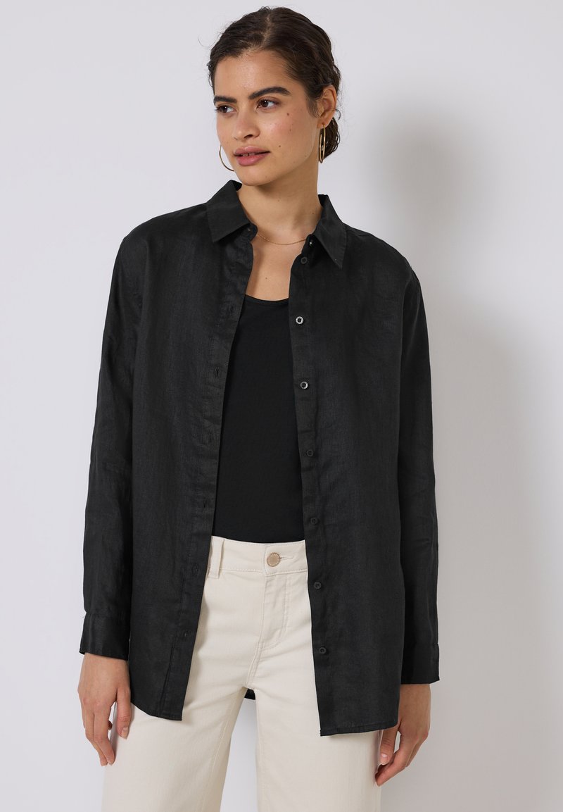 Camisa de lino negra con un frente abotonado, collar y mangas largas, combinada con una camiseta de tirantes negra y pantalones color crema. Textura suave.