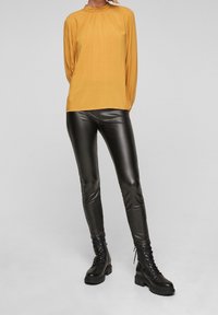 Senapsgul långärmad blus med veck, tillsammans med svarta glänsande imitationläderleggings och svarta snörade militärstövlar.