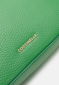 Portafoglio in pelle verde testurizzata con logo dorato "Coccinelle" sul davanti. Bordo liscio e design minimalista.