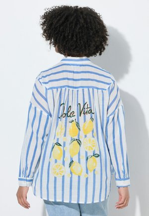 Osoba o kręconych włosach nosząca niebiesko-białą, paskowaną koszulę z żółtymi wzorami cytryn i napisem "Dolce Vita" na plecach.