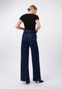 Sieviete ar gaišiem matiem, kas sakrauti bunā, ģērbjas melnā pieguļošā īsām piedurknēm blūzē, augsto jostasvietu platās kājās tumši zilās džinsās un melnos augstpapēžu apavos.