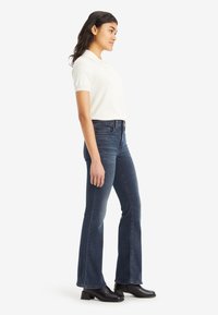 Slim fit jeans v temno denim barvi s širokim oprsjem, kombinirane s kratkosleeve polo majico v kremni barvi. Črne gležnjarje dopolnjujejo videz.