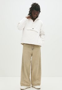 Junge Person mit Dreadlocks, die eine weiße Kapuzenjacke, beigen weiten Jeans und weiße Sneaker trägt und nach unten schaut, während eine Hand nahe am Kragen ist.
