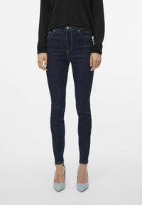 Tanke tamnoplave traperice s pojasom srednje visine, suptilnim žutim šavovima, dizajnom sa pet džepova, u paru s svijetloplavim cipelama na visoku petu sa šiljastim vrhom.