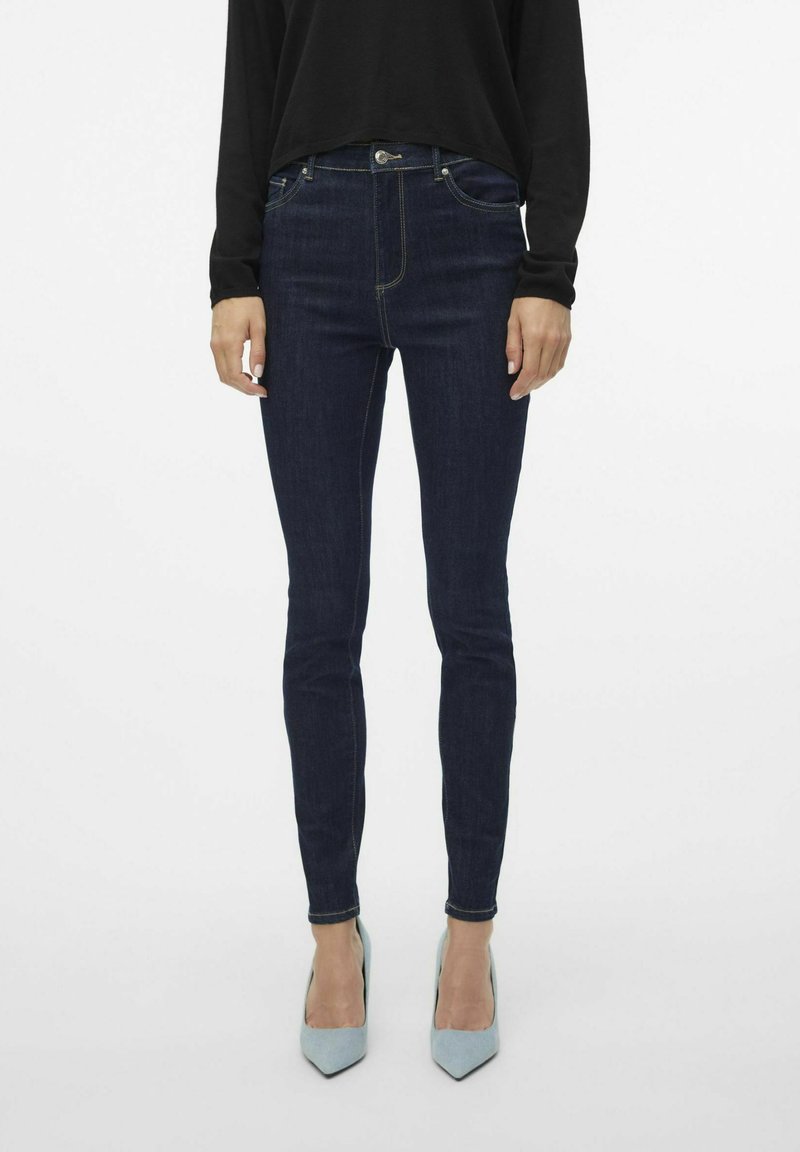 Tanke tamnoplave traperice s pojasom srednje visine, suptilnim žutim šavovima, dizajnom sa pet džepova, u paru s svijetloplavim cipelama na visoku petu sa šiljastim vrhom.