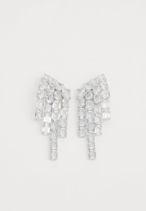 Przejrzyste kryształowe kolczyki o wydłużonym kształcie z prostokątnymi i kwadratowymi kamieniami kaskadowo układającymi się w kształcie wachlarza. Lśniąca struktura powierzchni.