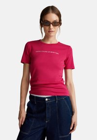 Fitted tričko s krátkým rukávem z vibranční růžové bavlny s bílým textem "UNITED COLORS OF BENETTON". Nosené s tmavě modrými denimovými kalhotami.