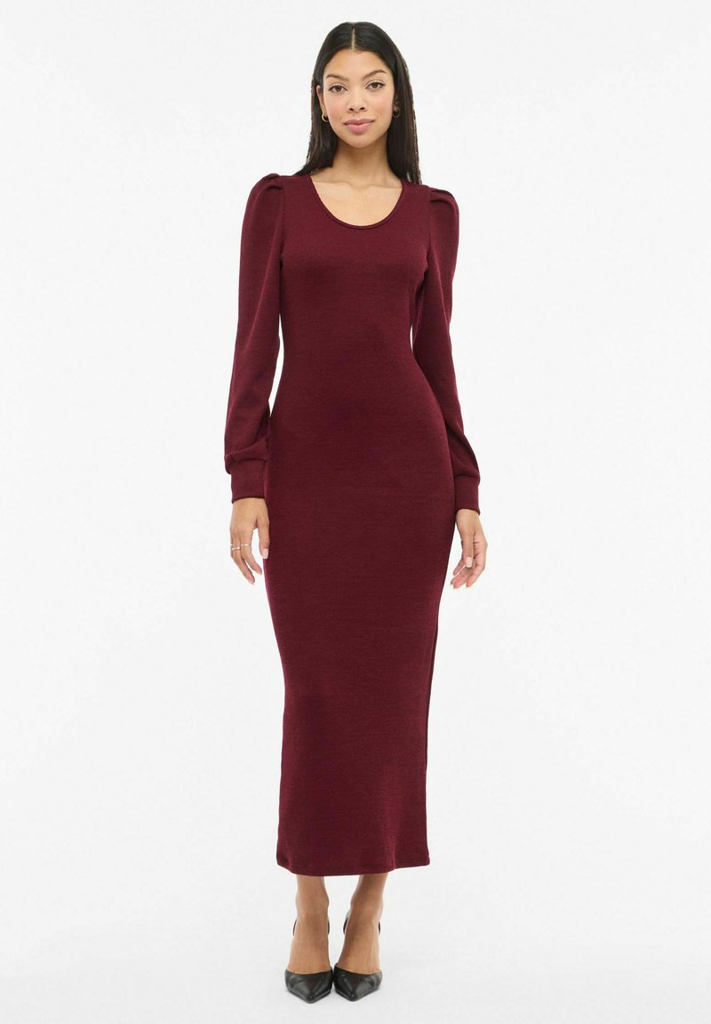 Mujer de pie vistiendo un vestido largo ajustado color burdeos con hombreras abullonadas y mangas largas, combinado con tacones negros puntiagudos.