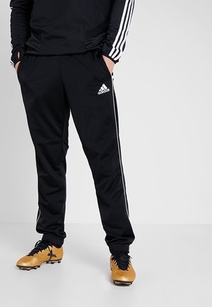 Osoba nosząca czarne spodnie dresowe Adidas z białymi paskami i logo, w połączeniu z żółto-czarnymi korkami piłkarskimi, ręce w kieszeniach.