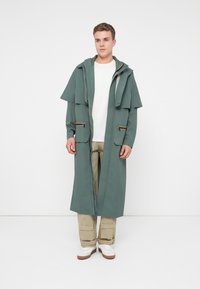 Νεαρός άνδρας που στέκεται φορώντας μακρύ πράσινο μπουφάν με κουκούλα, λευκή μπλούζα, μπεζ cargo παντελόνι και λευκά αθλητικά παπούτσια μπροστά σε λευκό φόντο.