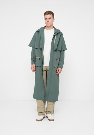 Jaunas vyras stovi ilgame žaliame gobtuvu paltas, baltame marškinėlyje, bežinėse krovinių kelnėse ir baltuose sportiniuose batuose prieš paprastą baltą foną.