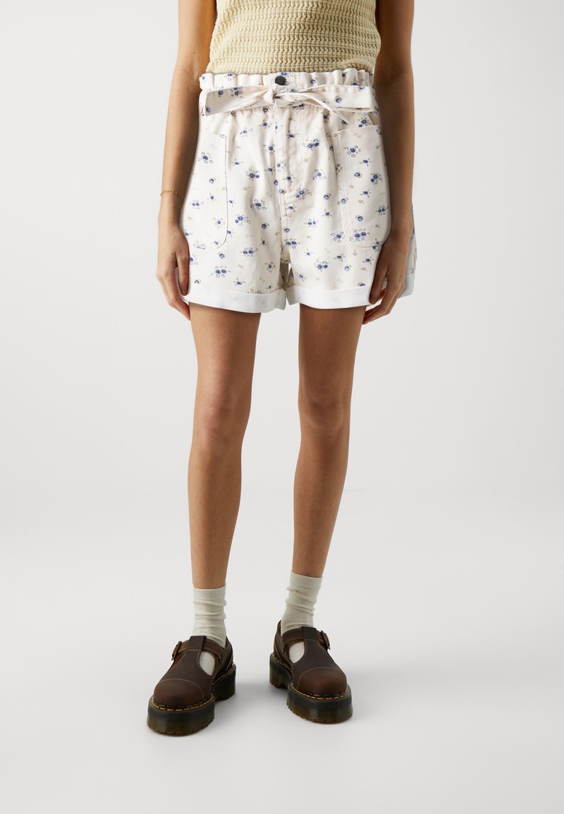 Pantaloni corti bianchi con vita arricciata e dettaglio di fiocco, dotati di tasche frontali e orli arrotolati. Indossati con scarpe marroni e calze verdi.