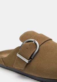 Brune ruskinds slide sandal med en bred rem med en metallisk spænde og en struktureret sort gummisål. Nærbillede fremhæver detaljer.