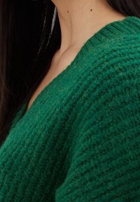 Zielony sweter dziany z głębokim dekoltem w kształcie litery V. Teksturowana tkanina wydaje się miękka i ciepła, z prążkowanym wykończeniem przy dekolcie.