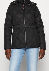 Svart quiltad pufferjacka med dragkedja fram, knäppdetaljer och cinchad midja. Har en huva och långa ärmar.