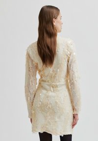 Kobieta z długimi brązowymi włosami, ubrana w teksturowaną sukienkę z kremowych cekinów i czarne rajstopy, stojąca tyłem na tle jasnego, jednolitego tła.