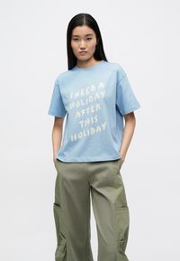 Ανοιχτό μπλε βαμβακερό t-shirt με κοντά μανίκια, με λευκό κείμενο: «ΧΡΕΙΑΖΟΜΑΙ ΔΙΑΚΟΠΕΣ ΜΕΤΑ ΑΠΟ ΑΥΤΕΣ ΤΙΣ ΔΙΑΚΟΠΕΣ.» Συνδυασμένο με χακί cargo παντελόνια.