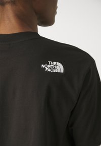 Zwarte katoenen t-shirt met een klein wit geborduurd logo van "The North Face" op de bovenrug, nauwsluitend ontwerp, gladde textuur.