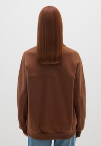 Persona con capelli rossi lisci di lunghezza media, che indossa una felpa marrone e jeans blu, in piedi di fronte a un muro bianco.