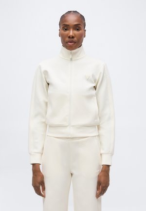 Femme aux cheveux tressés portant une veste blanche zippée et un pantalon assorti, debout devant un fond blanc uni.