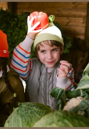 Bambino con cappello verde tiene un peperone rosso sulla testa, sorridendo tra verdure fresche, tra cui cavolo e cavolfiore.