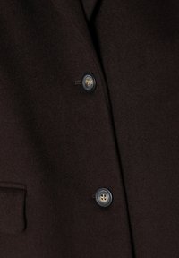Bruine wollen blazer met twee knopen, waarvan één zwart met een doorzichtig middenstuk, en een platte zak aan de linkerkant. Soepele textuur en een getailleerd ontwerp.