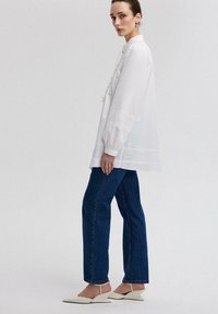 Camicia bianca button-up con maniche lunghe e cuciture decorative, abbinata a jeans dritti blu scuro e scarpe slingback bianche.