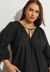 Svart långärmad blus med V-ringning och knytdetalj. Har texturerad tyg med blommig broderi. Rundade ärmar med elastiska manschetter.