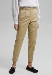 Pantaloni beige su misura con gamba dritta e dettagli delle pieghe. Realizzati in tessuto liscio, presentano un frontale piatto e tasche laterali, abbinati a mocassini blu navy.