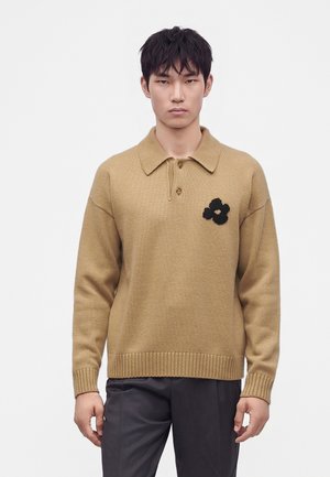 Junger Mann mit kurzen schwarzen Haaren, der einen beigen Strickpullover mit Kragen und einem schwarzen Blumenmotiv auf der Brust trägt, kombiniert mit dunkler Hose.