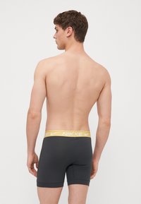 Svarta stretchboxers med en gul Nike-logga i midjan. Slät textur och figurnära design. Bilden visar plagget bakifrån.