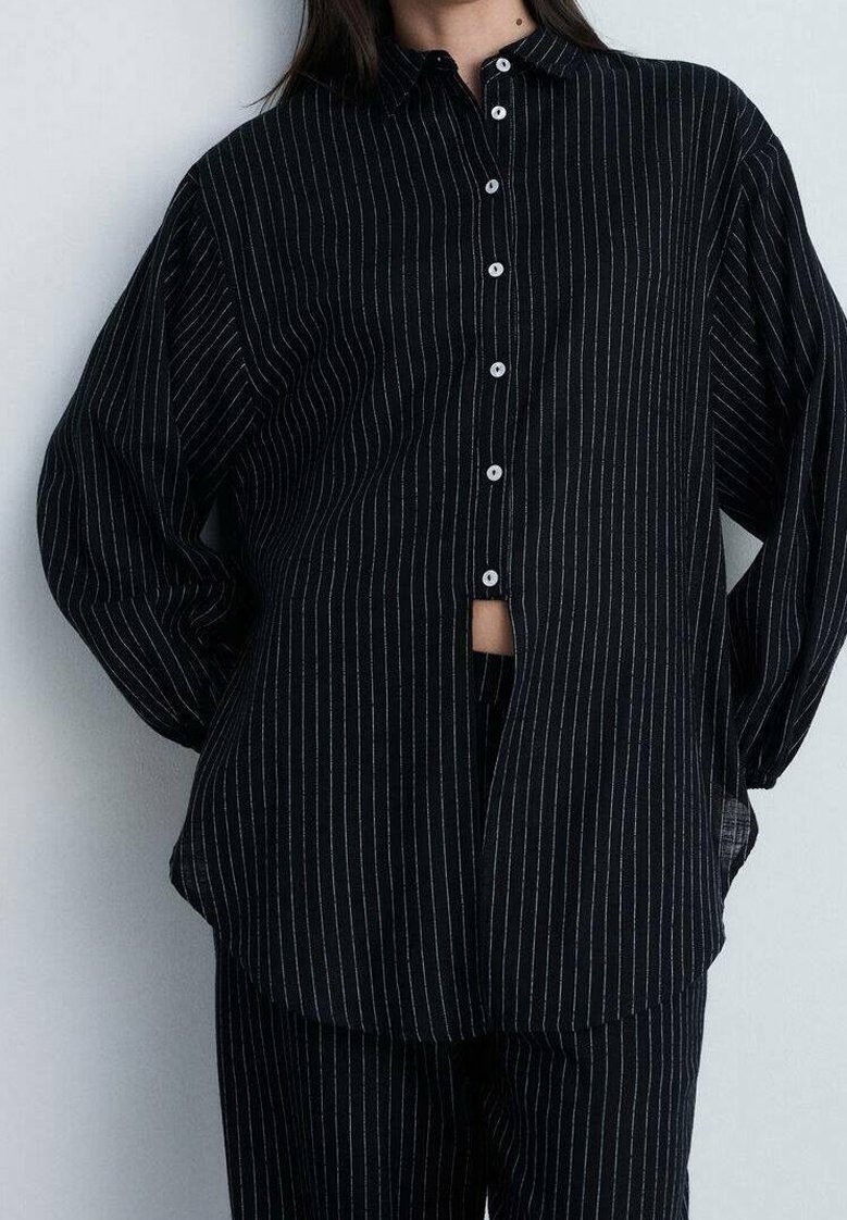 Personne portant une chemise noire à fines rayures avec des boutons blancs et un pantalon assorti à fines rayures, debout devant un fond uni.