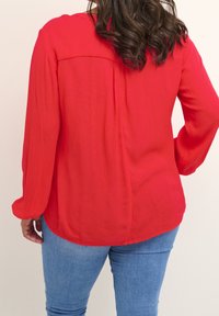 Blouse rouge à manches longues avec un tissu texturé, fentes latérales, un pli dans le dos et un ourlet arrondi, assortie à un jean bleu.
