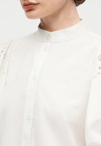 Κοντινό πλάνο ενός ατόμου που φοράει λευκή μπλούζα με ψηλό γιακά και κουμπιά στο μπροστινό μέρος, με λεπτομέρειες από δαντέλα με ανοίγματα στα μανίκια.