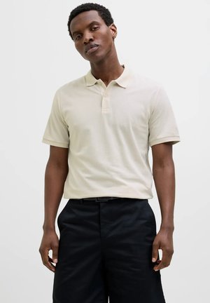 Polo shirt corto a maniche corte color beige chiaro con tessuto testurizzato, colletto e pattina frontale. Indossato con pantaloni corti neri su misura.