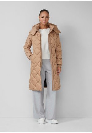 Manteau long beige matelassé avec capuche détachable, comprenant une fermeture éclair et des boutons-pression, associé à un pantalon large gris clair.