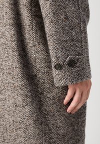 Palton cu model herringbone de culoare maro, realizat dintr-un material texturat din lână, având două butoane negre pe manșeta mâinii. Prim-plan al zonei brațului.