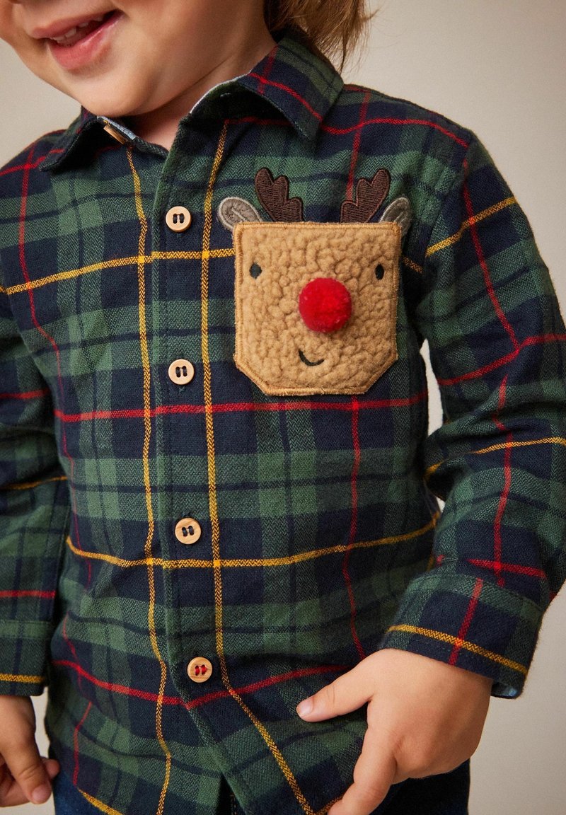 Enfant portant une chemise à carreaux verte et bleu marine avec un patch visage de renne avec un nez pompon rouge sur la poche poitrine.