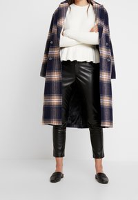 Pitkä ruudullinen takki laivastonsinisessä, tanin ja kerman sävyissä valkoisen ribbineuleen päällä, yhdistetty mustiin keinonahkahousuihin ja teksturoituihin mustiin loafereihin.