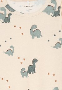 Beżowa odzież dziecięca z niebiesko-zielonymi nadrukami dinozaurów i rozsypanymi brązowymi odciskami dinozaurów.