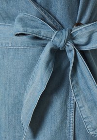 Peça de vestuário em denim azul claro com um cinto de laço. O tecido parece suave, com uma leve textura. Apresenta um detalhe em botão próximo ao nó.